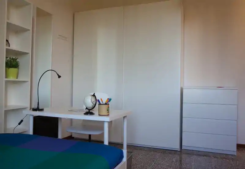 Bright, minimal bedroom with a white wardrobe and chest of drawers. The image shows a bed with a colorful cover in the foreground, a tidy desk with a globe and pencils, and simple Scandinavian-style furnishings.