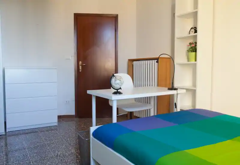 Bright, tidy bedroom with a colorful double bed in the foreground, a white desk and chair, shelving and a chest of drawers — suitable for a student or single occupant.