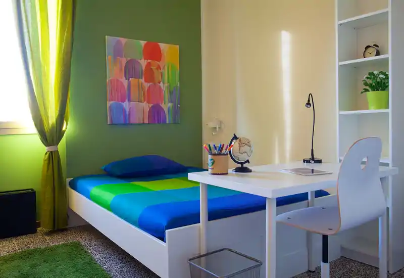 Camera singola luminosa e colorata con letto ordinato, scrivania e scaffalatura — ideale per uno studente o una persona sola. Luce naturale e decor vivaci creano uno spazio accogliente per dormire e lavorare.