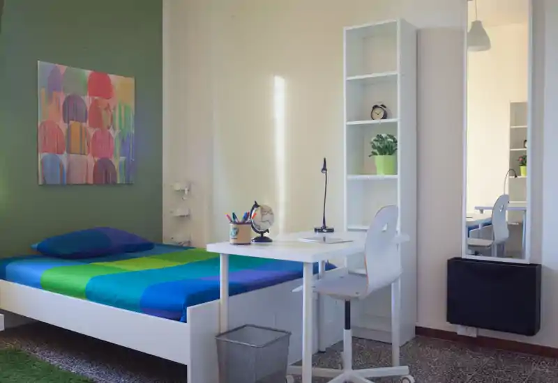 Bright, tidy bedroom with a colorful bed and a compact study area: white bed frame with blue-green linens, bedside artwork, desk with lamp and shelving — ideal for students or a single occupant.