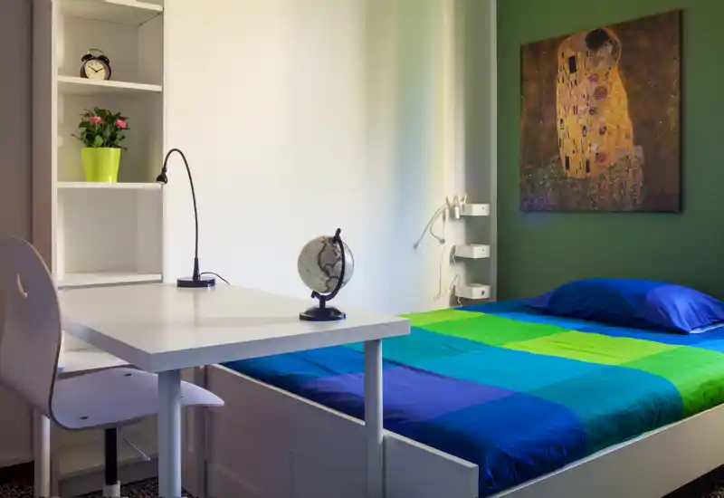 Bright, neatly styled single bedroom with a colorful blue-green bedspread, compact desk with lamp and globe, shelving and decorative artwork — ideal for a student or short-term rental.