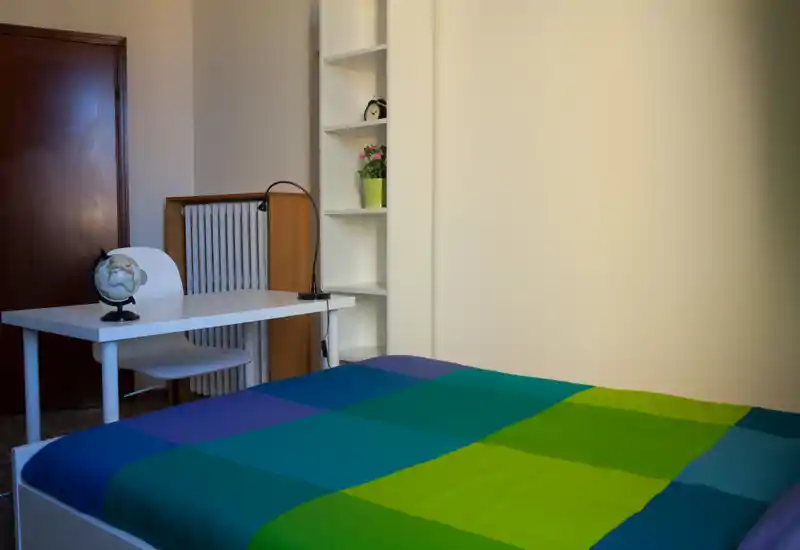 Bright, tidy bedroom with a colorful queen-size bed in the foreground, a small desk and chair at the side, and shelving with decorative accents — ideal for listings highlighting a cozy private room.