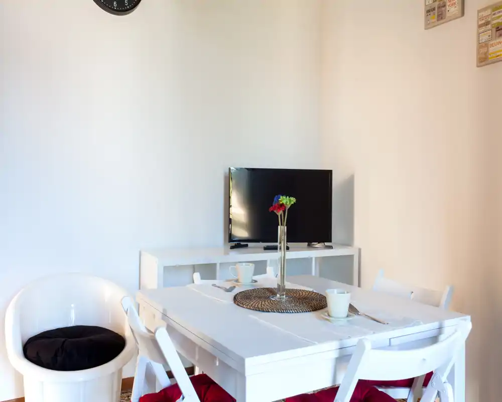 Bright, minimalist dining area with a white table set for four, decorative vase with flowers and a TV console in the background — ideal for compact apartment listings.