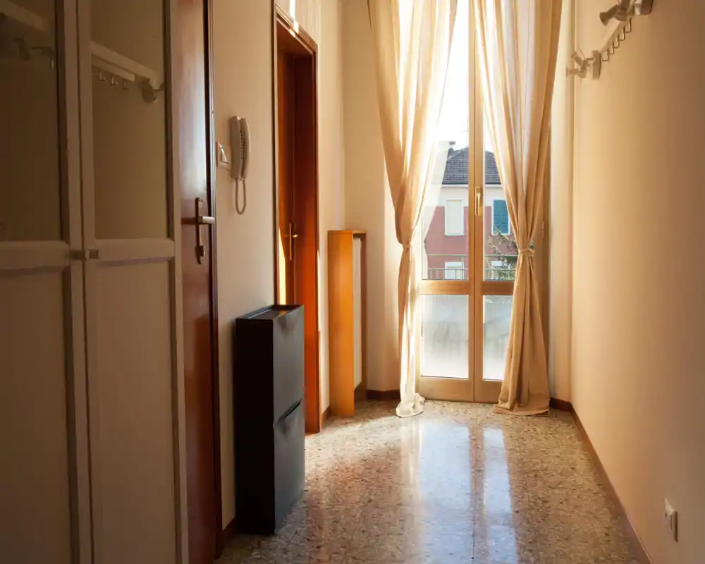 Bright entrance hallway with large glass door, soft curtains and terrazzo floor; compact storage units and wall hooks visible, ideal for welcoming guests.