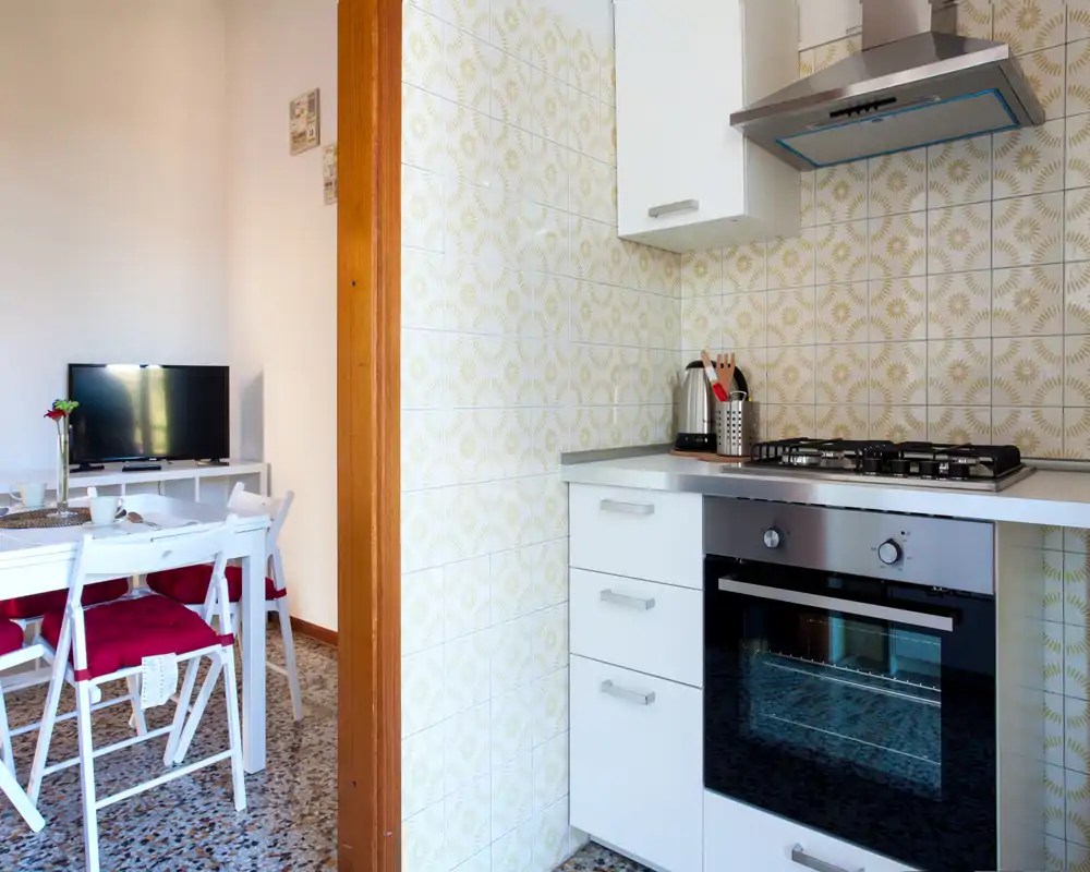 Bright, compact kitchen with ceramic-tiled backsplash, gas hob and built-in oven; adjacent dining area with white table and chairs visible, creating a practical and welcoming space.