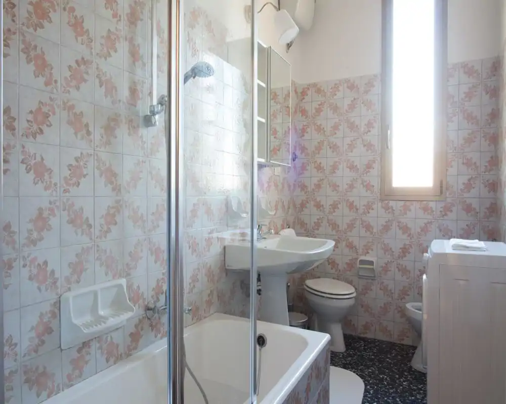 Bright, vintage bathroom with floral ceramic tiles, a bathtub with glass screen, pedestal sink, toilet and a window providing natural light.