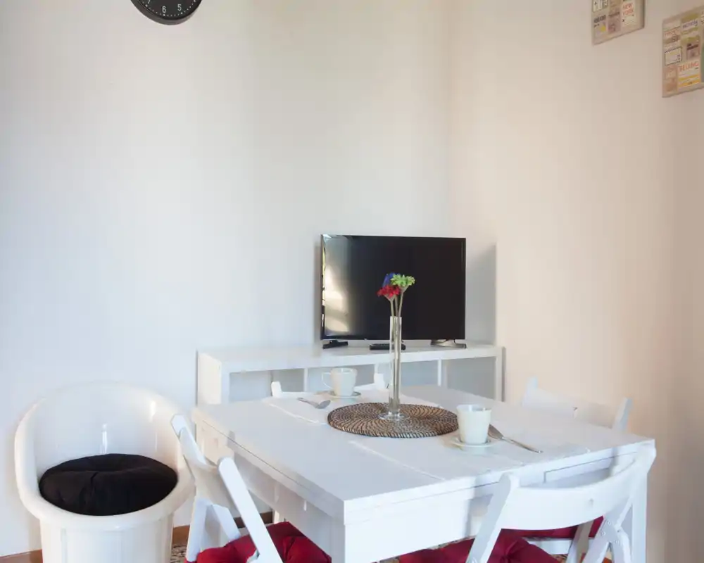 Bright, minimalist dining area with a white table set for two, red-cushioned folding chairs and a small vase centerpiece; TV and side console visible in the background.