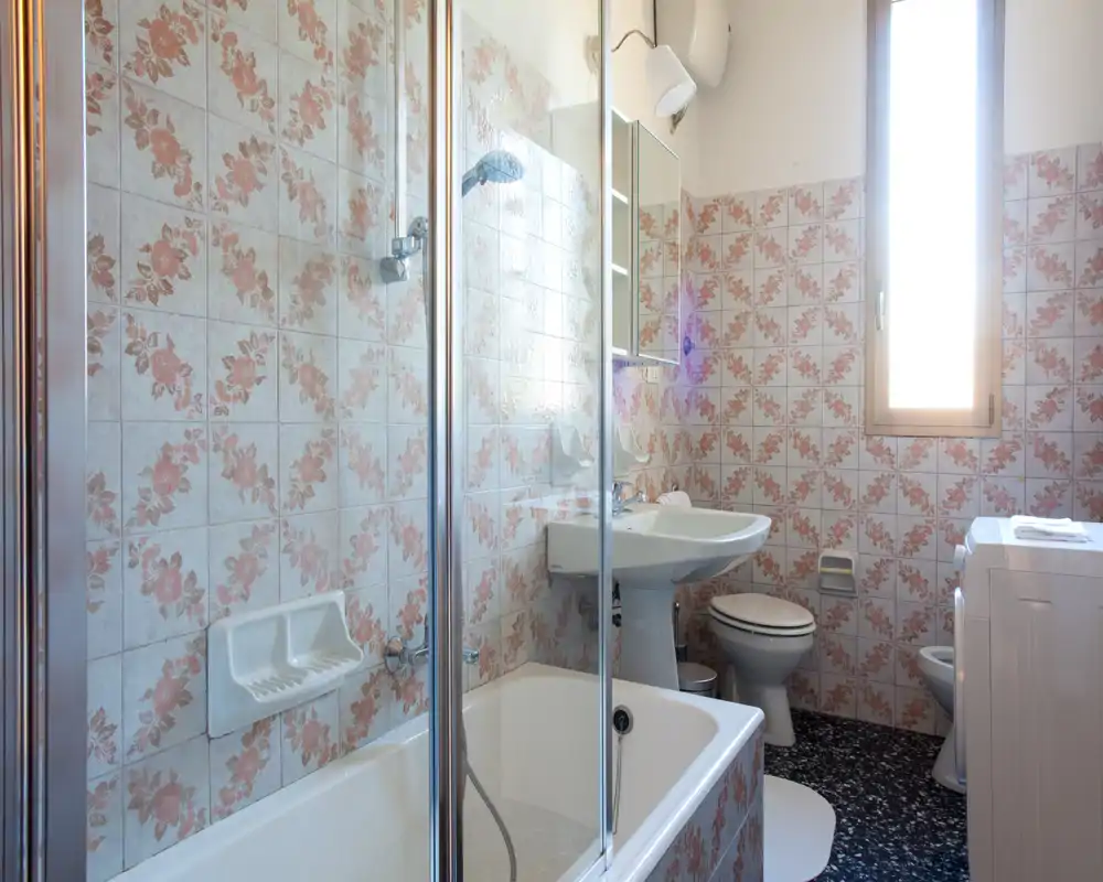 Bright, well-kept bathroom with bathtub and glass shower screen, pedestal sink, toilet and patterned wall tiles. Natural light enters through a tall window.