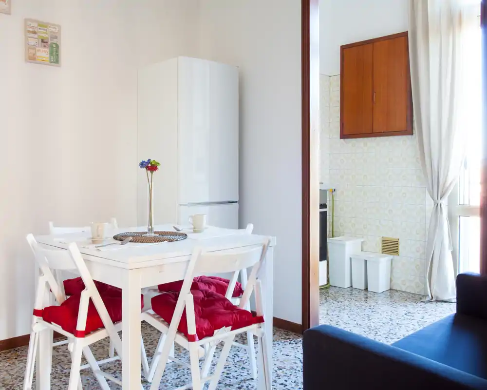 Bright, cozy living/dining corner with a small white table, four chairs with red cushions, and a refrigerator in the background — ideal for a compact apartment.