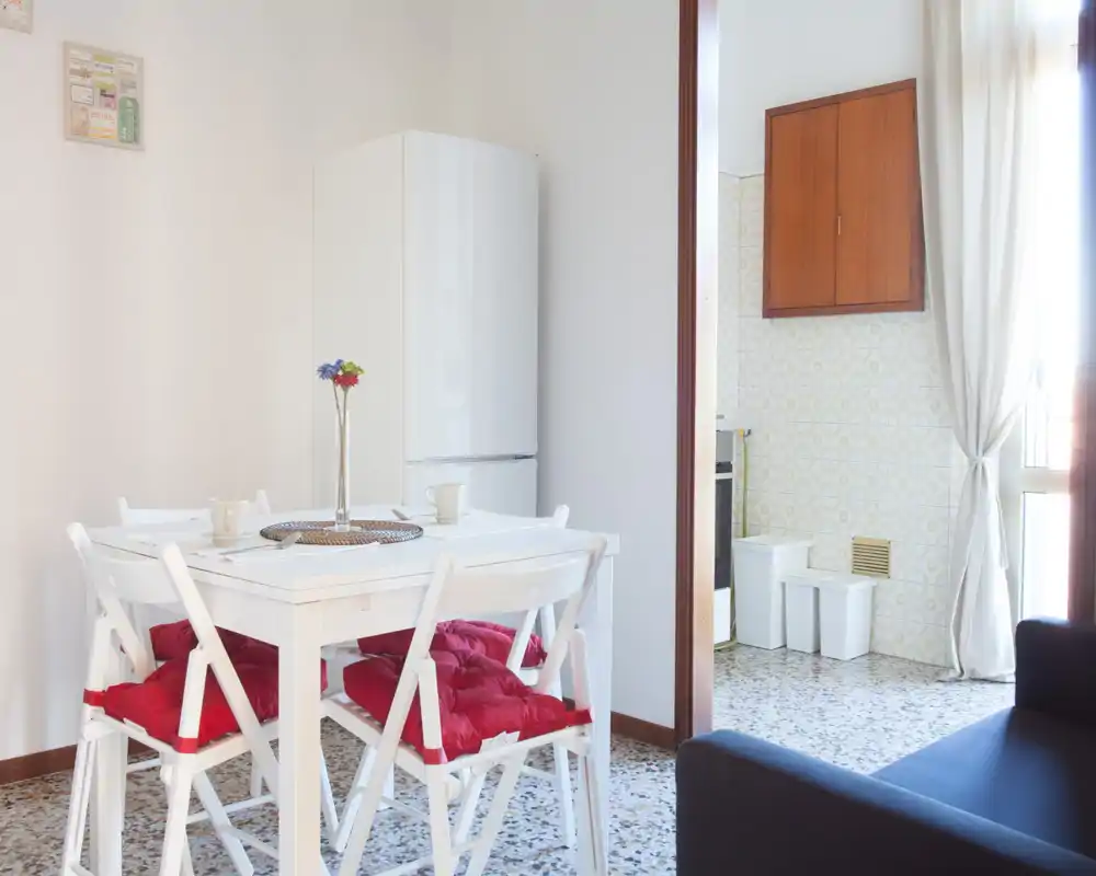 Bright dining nook with a white table and four cushioned chairs, adjacent to a small kitchen and large window—simple, airy and welcoming.