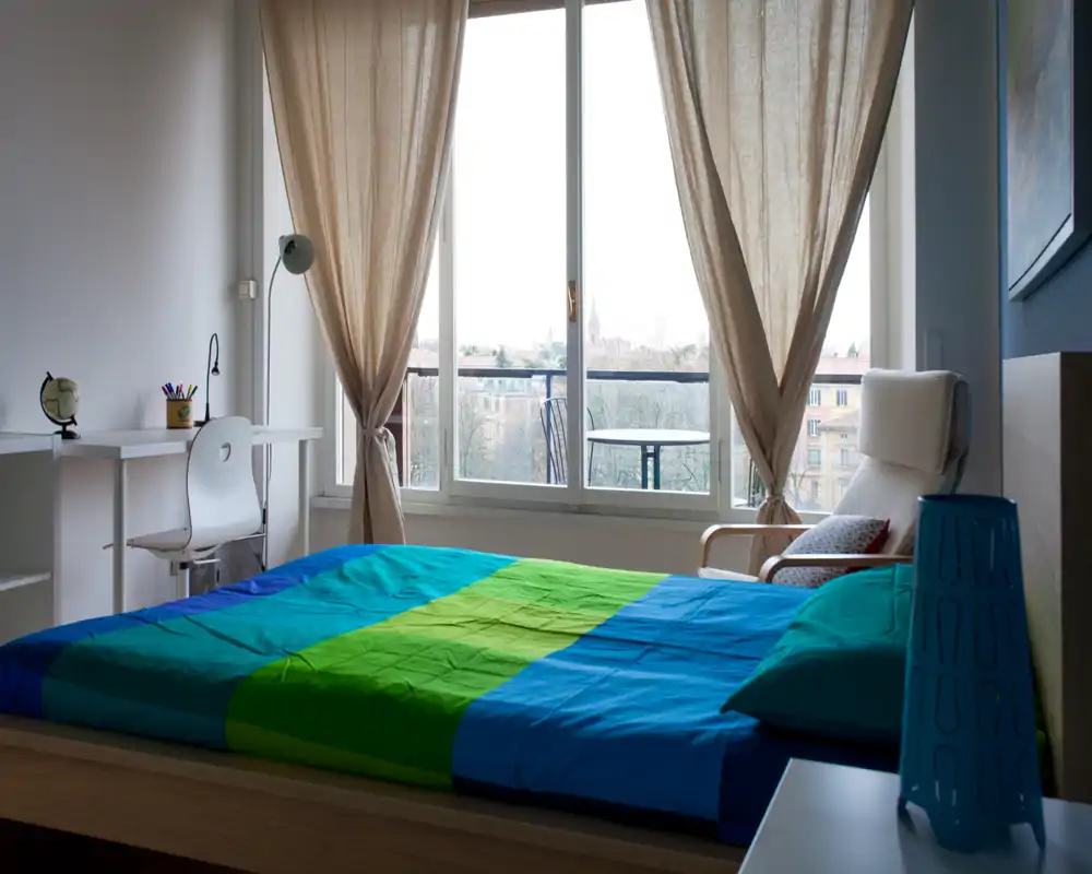 Chambre lumineuse et soignée avec grande fenêtre et vue sur balcon ; couette colorée et fauteuil confortable créent une ambiance chaleureuse.