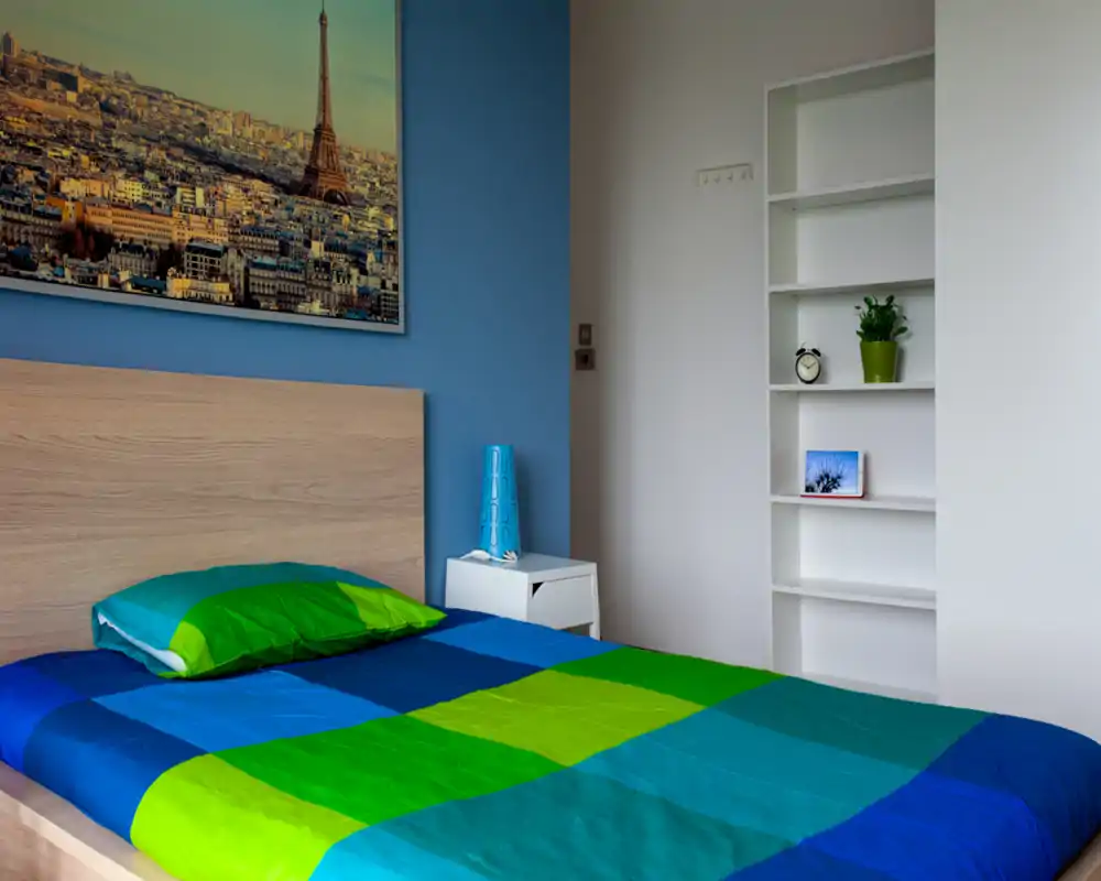 Chambre lumineuse et colorée avec tête de lit en bois, couette bleu-vert à motifs et table de chevet avec lampe décorative. Des étagères et une photo de ville complètent cet espace moderne et chaleureux.