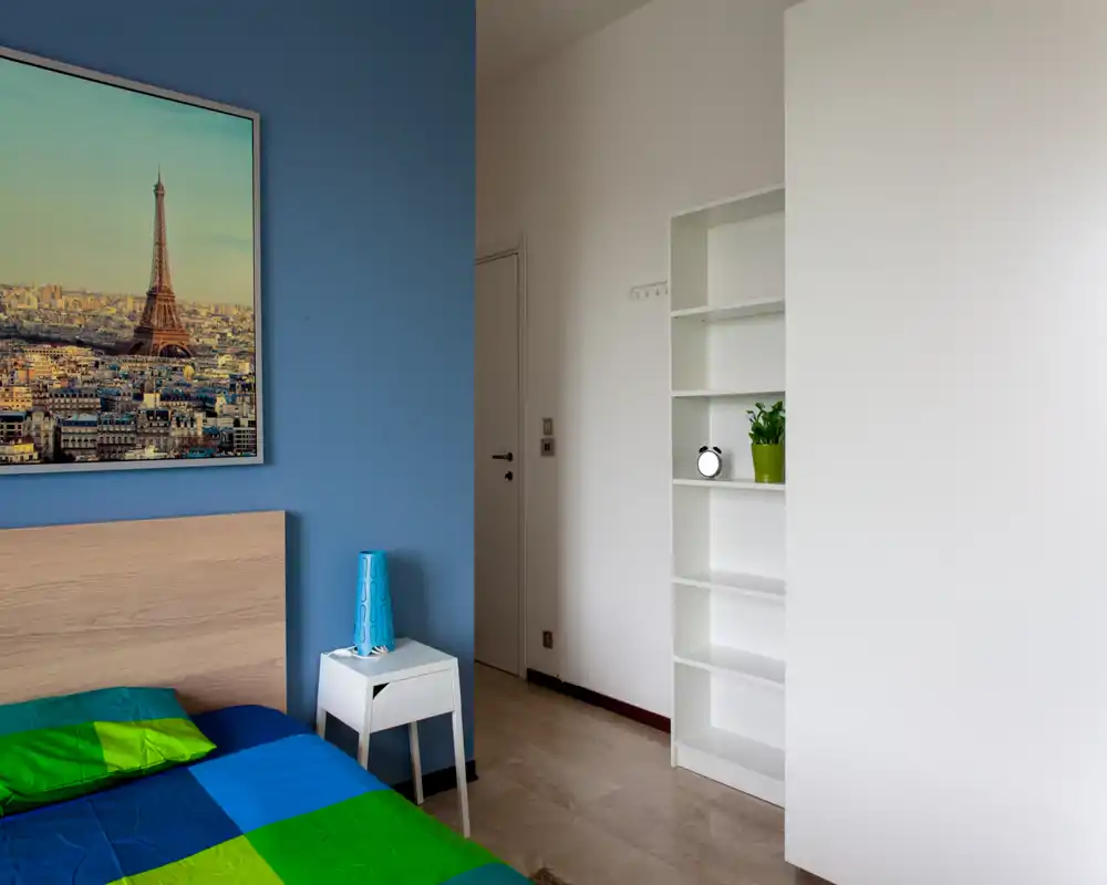 Chambre moderne et lumineuse avec mur d'accent bleu, tête de lit en bois et linge de lit coloré. Étagères épurées et lumière naturelle offrent une atmosphère aérée, idéale pour une annonce locative.