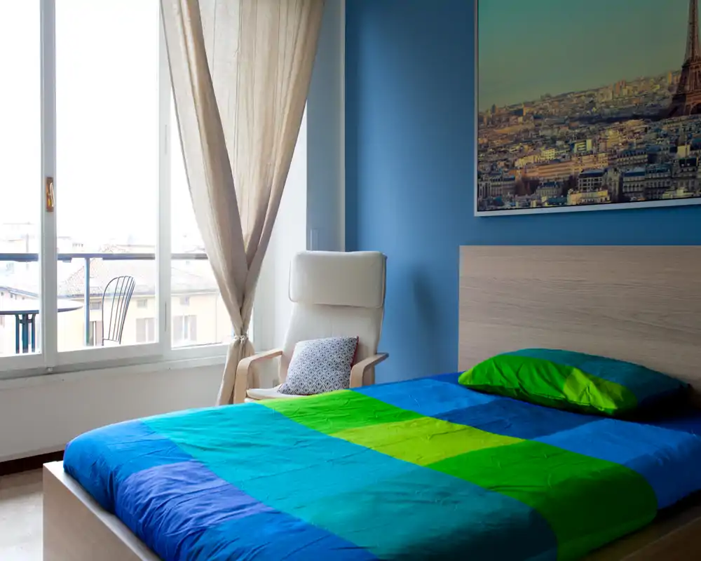 Chambre moderne et lumineuse avec grande fenêtre, lit aux draps colorés et fauteuil relax près de la fenêtre donnant sur un petit balcon.