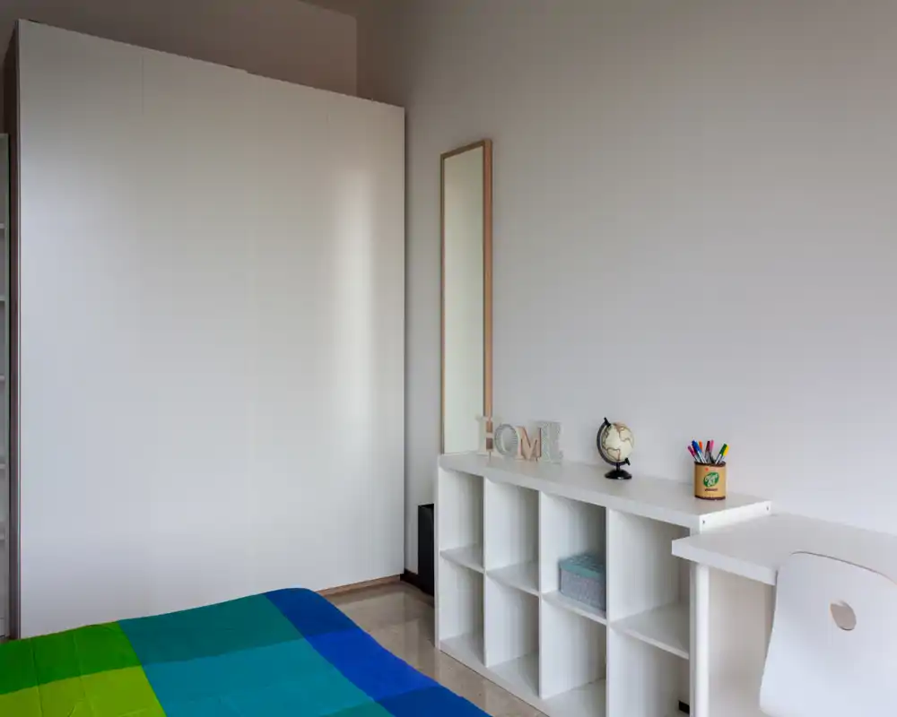 Chambre lumineuse et minimaliste avec une couette colorée, une armoire blanche et des étagères; un petit coin bureau avec chaise et objets décoratifs crée un espace de nuit moderne et soigné.