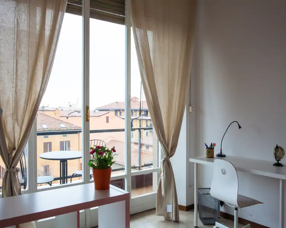 Habitación luminosa con gran ventana de suelo a techo y balcón, con un pequeño escritorio, silla y una planta decorativa; las cortinas neutras aportan calidez.