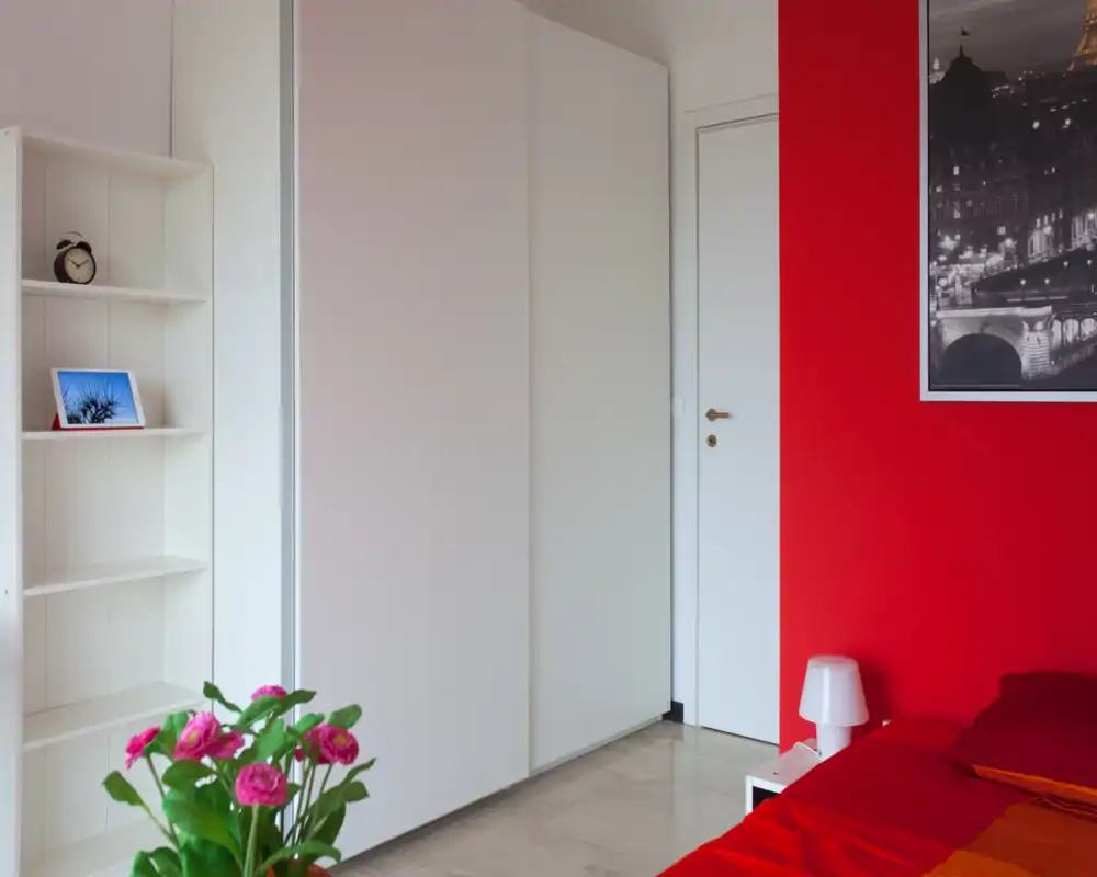 Rincón de habitación ordenado con pared roja, lámpara de mesita y cama parcialmente visible con ropa de cama cálida; armario empotrado y estanterías ofrecen almacenamiento y un aire moderno y luminoso.