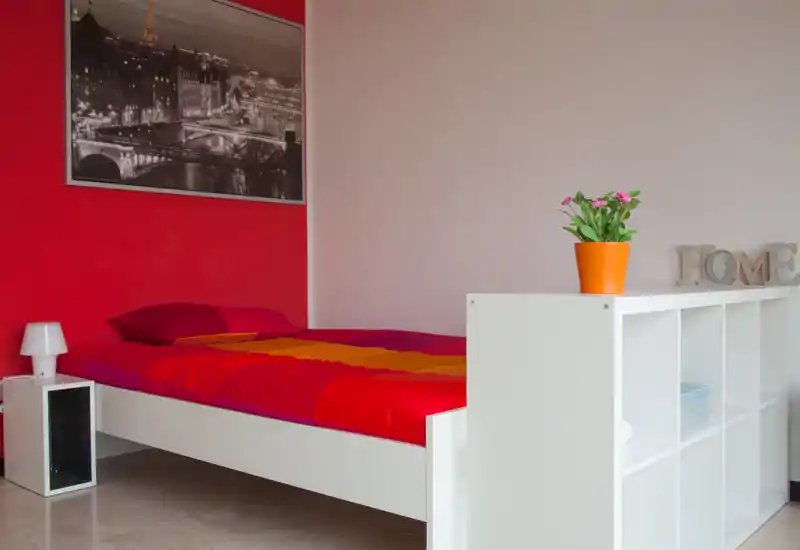 Dormitorio moderno y luminoso con una cama doble de colores vibrantes contra una pared roja, lámpara de noche sencilla y módulo blanco de almacenamiento que aportan orden y estilo contemporáneo.
