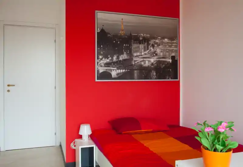 Dormitorio luminoso y colorido con cama doble, ropa de cama rojo-naranja, mesita y cuadro decorativo de paisaje urbano. Una planta en maceta añade un toque fresco.