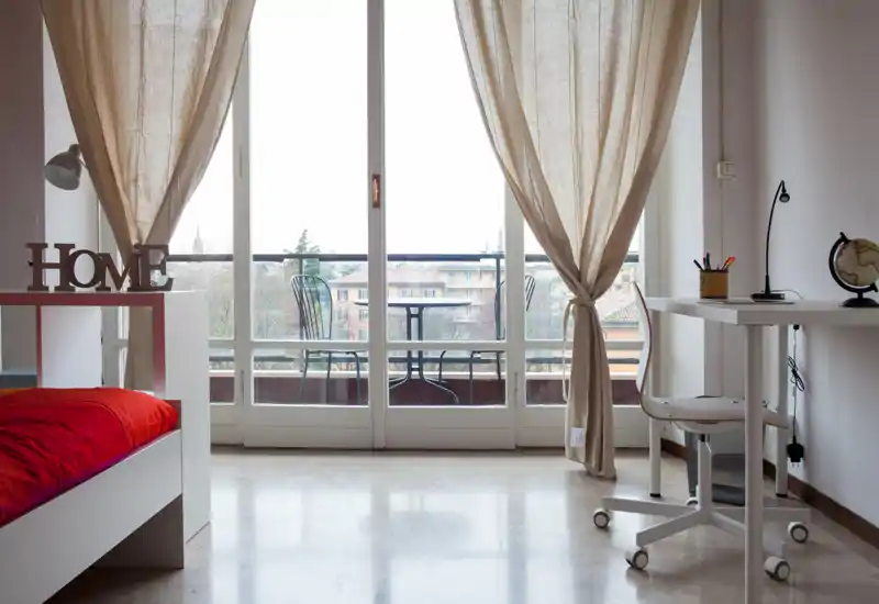 Dormitorio luminoso y minimalista con cama individual de ropa de cama roja, un escritorio y grandes puertas al balcón que llenan la habitación de luz natural.