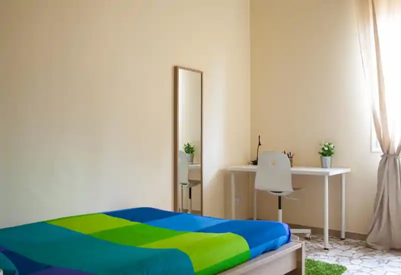 Sunny, minimalist bedroom with a colorful bedspread, a simple desk and chair by the window, and a full-length mirror — bright and tidy space ideal for short stays.