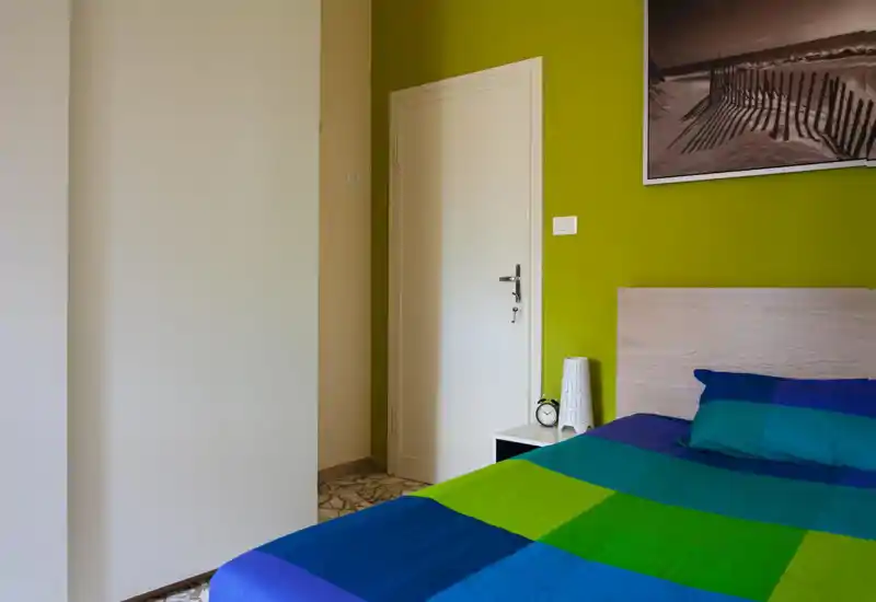 Bright, modern bedroom corner with a wooden headboard, colorful blue-green bedding and a bedside table with lamp and alarm clock — ideal for a cozy city stay.