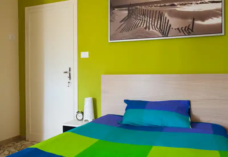 Bright, modern bedroom with colorful bedding, wooden headboard and a bedside table with lamp and clock. The room features a vibrant green accent wall and framed artwork above the bed.