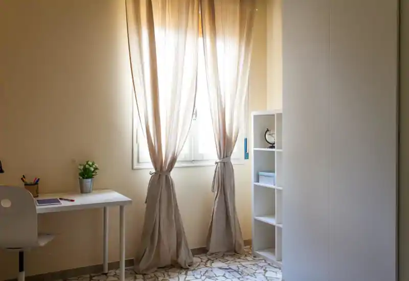 Bright minimal bedroom corner with soft beige curtains, a small white desk and shelving; ideal for a calm workspace near the window.