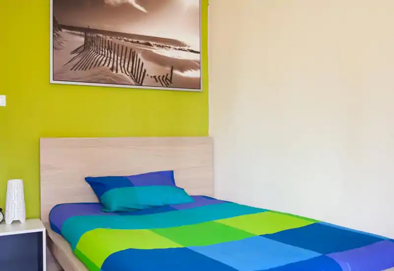 Bright, modern bedroom with a colorful blue‑and‑green bedspread, visible headboard and a seaside framed print on a lime accent wall.