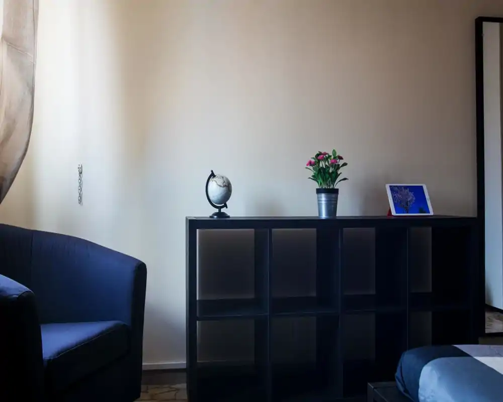 Calm, simply styled bedroom corner with a small visible portion of the bed, a dark shelving unit topped with a globe, a potted plant and a tablet — soft natural light creates a relaxed atmosphere.