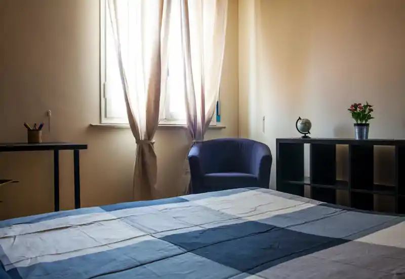 Bright, simply styled bedroom with a large bed in the foreground, soft natural light from the window, a cozy armchair and minimalist shelving.