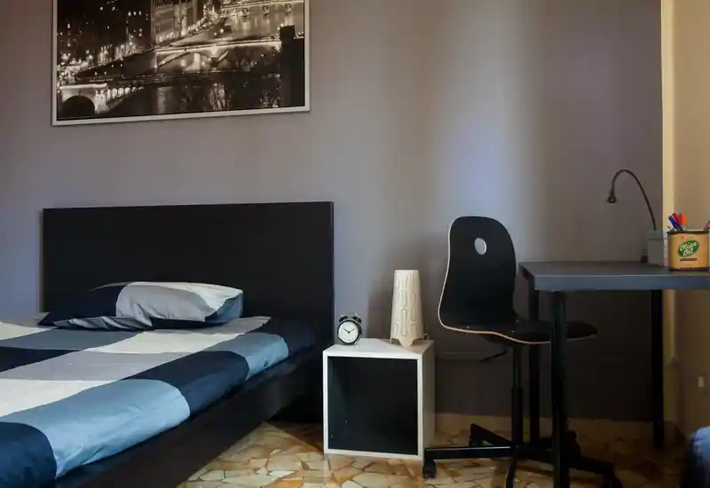 A neat bedroom with a large bed in the foreground, simple bedside table with an alarm clock and a desk with chair—bright, tidy and suitable for a rental listing.