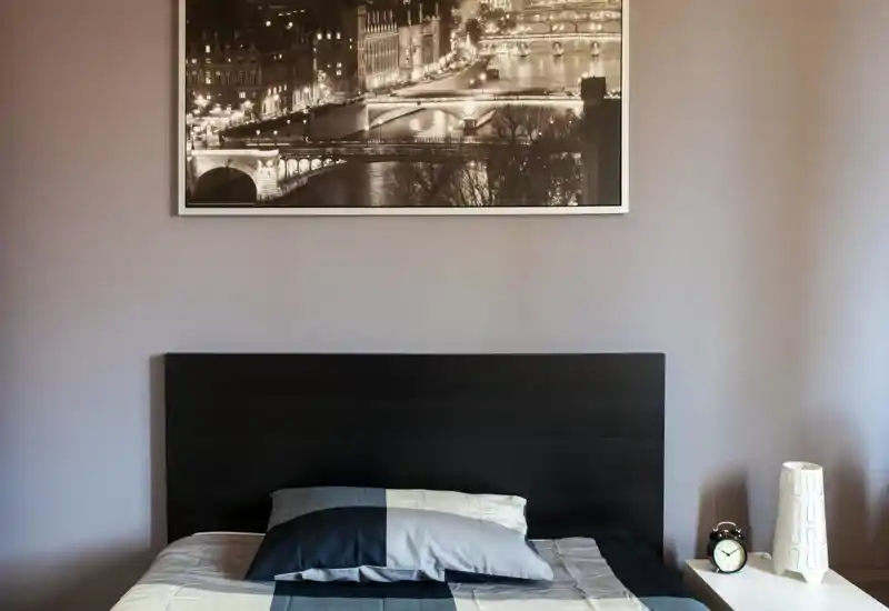 Modern bedroom with a black headboard and a neatly made bed in neutral blue-gray bedding; a framed city photo hangs above and a bedside table holds an alarm clock and decorative lamp.