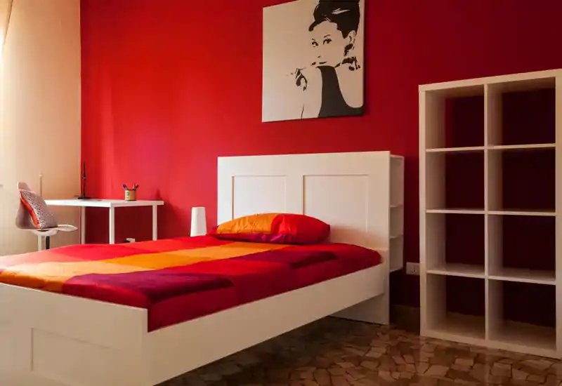 Dormitorio moderno y luminoso con una cama de estructura blanca, ropa de cama roja y naranja y un escritorio minimalista. Pared de acento roja y estantería blanca que aportan un ambiente elegante y acogedor.