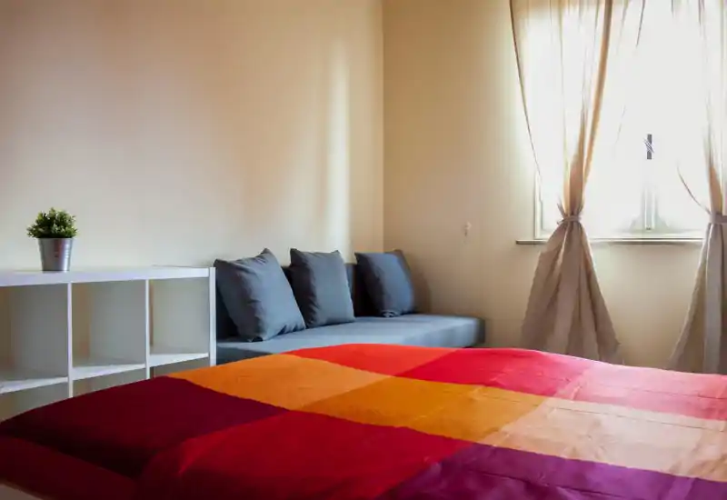 Dormitorio luminoso y acogedor con un amplio edredón colorido en primer plano, un sofá gris con cojines junto a la pared y suave luz natural desde la ventana.