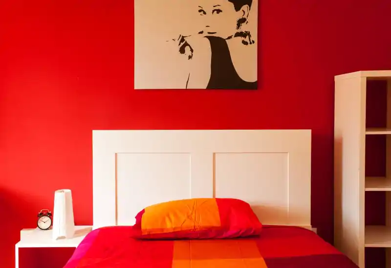 Dormitorio luminoso y con estilo con cabecero blanco sobre pared roja intensa, ropa de cama rojo-naranja y decoración de mesita sencilla — ideal para un anuncio moderno.