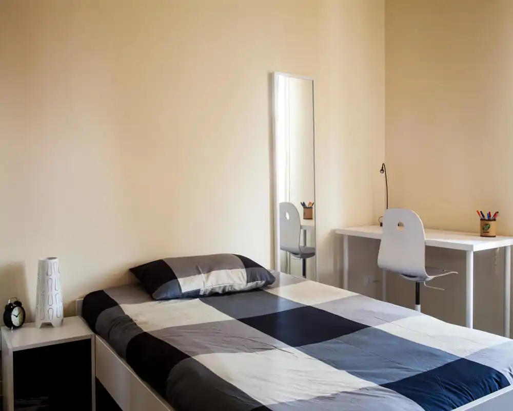 Camera da letto luminosa e minimal con letto matrimoniale ordinato in toni neutri, comodino con lampada e sveglia e una scrivania semplice con sedia — perfetta per un affitto moderno e tranquillo.