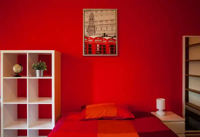 Dormitorio luminoso y con estilo, pared de acento roja y cama bien hecha con ropa de cama rojo-anaranjada; estantería sencilla y mesita con lámpara completan el conjunto.
