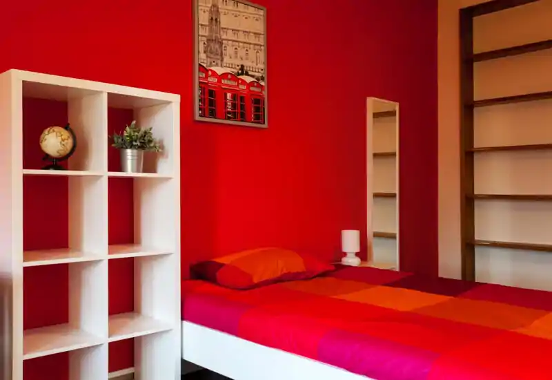 Dormitorio moderno y luminoso con pared roja y cama doble con ropa de cama roja-anaranjada; estantería blanca y espejo aportan almacenamiento y reflejan la luz para un ambiente acogedor y con estilo.