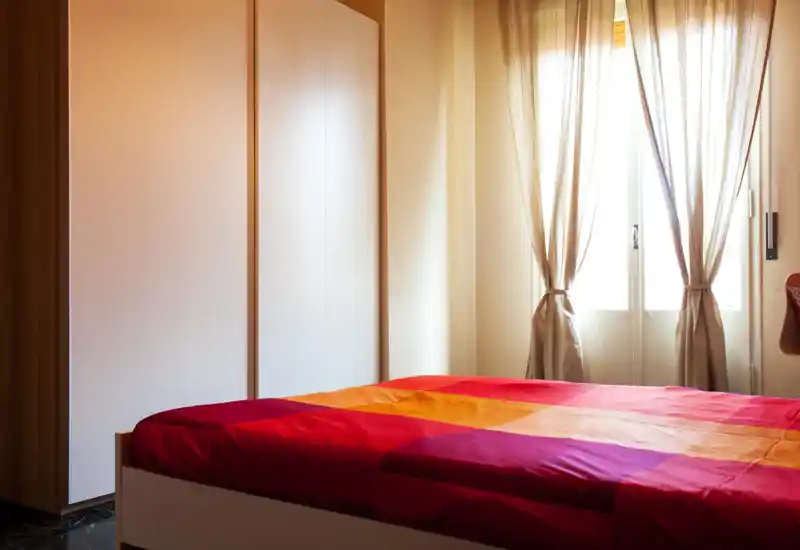 Dormitorio luminoso y acogedor con una colcha colorida y luz natural suave desde una ventana con cortinas anudadas. Un gran armario y tonos cálidos generan una atmósfera agradable.