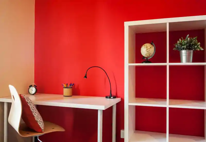 Rincón de escritorio moderno y luminoso con mesa blanca, silla y estantería frente a una pared roja intensa — ideal para oficina en casa o zona de estudio.