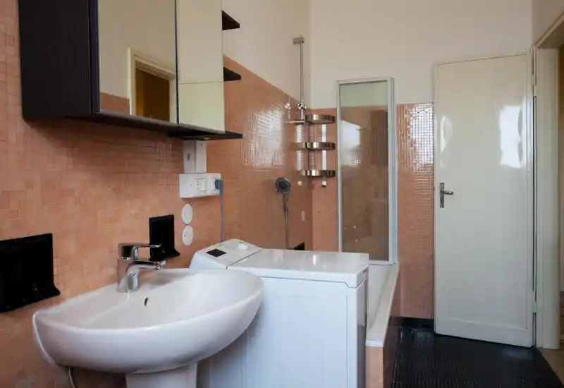Bagno compatto con piastrelle mosaico color pesca, lavabo bianco, lavatrice accanto alla vasca e pannello doccia in vetro — ordinato e ben illuminato.