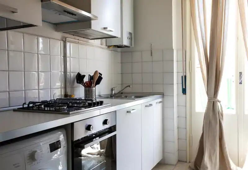 Cucina compatta e luminosa con piano cottura a gas, forno integrato, lavello in acciaio e utensili disposti ordinatamente accanto alla porta finestra che illumina l'ambiente.
