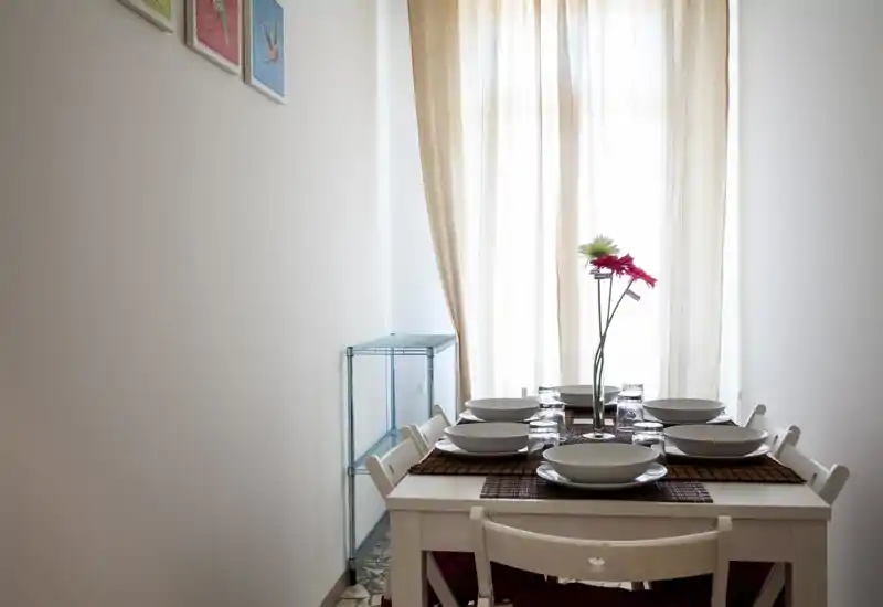 Tavolo da pranzo ben apparecchiato per sei persone con stoviglie bianche e un vaso di fiori vicino a una finestra luminosa — un angolo pranzo minimal e accogliente.