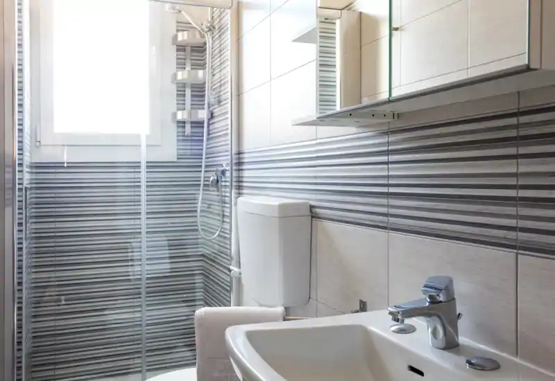 Bagno moderno e ben illuminato con box doccia in vetro, cassetta wc a muro, lavabo e rubinetteria cromata contemporanea: piastrelle grigie a strisce donano un aspetto pulito e di design.
