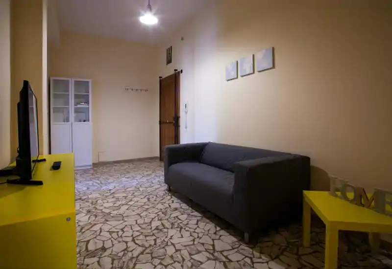Salón sencillo y bien iluminado con sofá gris, mueble de TV amarillo y mesa auxiliar a juego; decoración minimalista y suelo de baldosas que crean un espacio limpio y funcional.