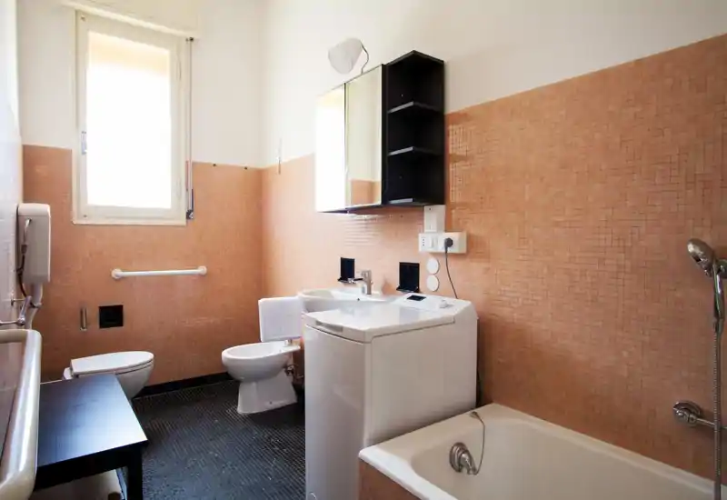 Bagno luminoso e funzionale con vasca, bidet, WC e lavabo; piastrelle mosaico color pesca e una lavatrice compatta per maggiore praticità.