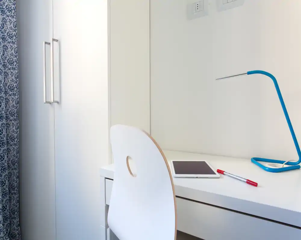 Angolo di camera luminoso e compatto con scrivania bianca, sedia moderna e lampada da tavolo blu; la tenda decorata e l'armadio suggeriscono un'area notte funzionale.
