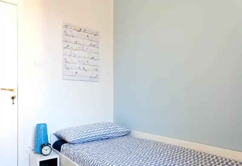 Camera singola luminosa e ordinata con letto con copertura a motivo, comodino e parete accentuata azzurra: ideale per un soggiorno confortevole.