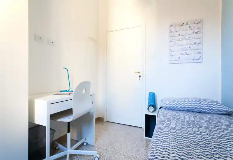 Camera singola compatta e luminosa con letto dal motivo geometrico, comodino a colonna e una scrivania funzionale ideale per lo studio. Arredamento minimale e luce naturale creano un'atmosfera tranquilla e accogliente.